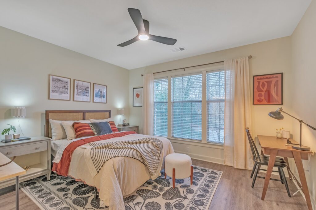 A cozy bedroom with a large window, a bed with striped and patterned bedding, two side tables with lamps, wall art above the bed, a desk with a chair and lamp, and a modern ceiling fan.