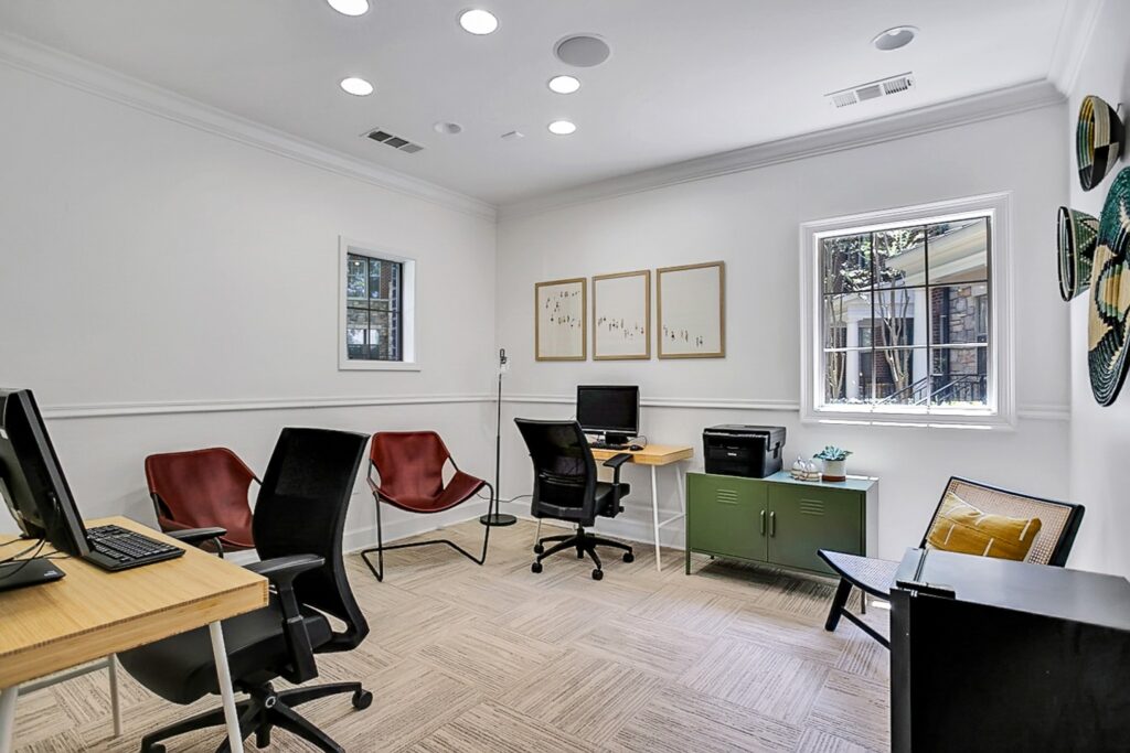 A modern office space with three red chairs, two desks with computers, framed art on white walls, a window, and a green cabinet with a printer. Decorative plates hang on one wall, and a yellow pillow sits on a chair.