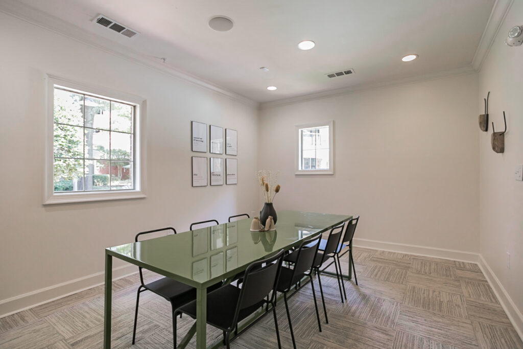A minimalist meeting room with a green table, eight black chairs, a vase with dried flowers, two windows, framed art on the wall, and antler decor. The space is bright and modern with neutral-colored walls and carpet.