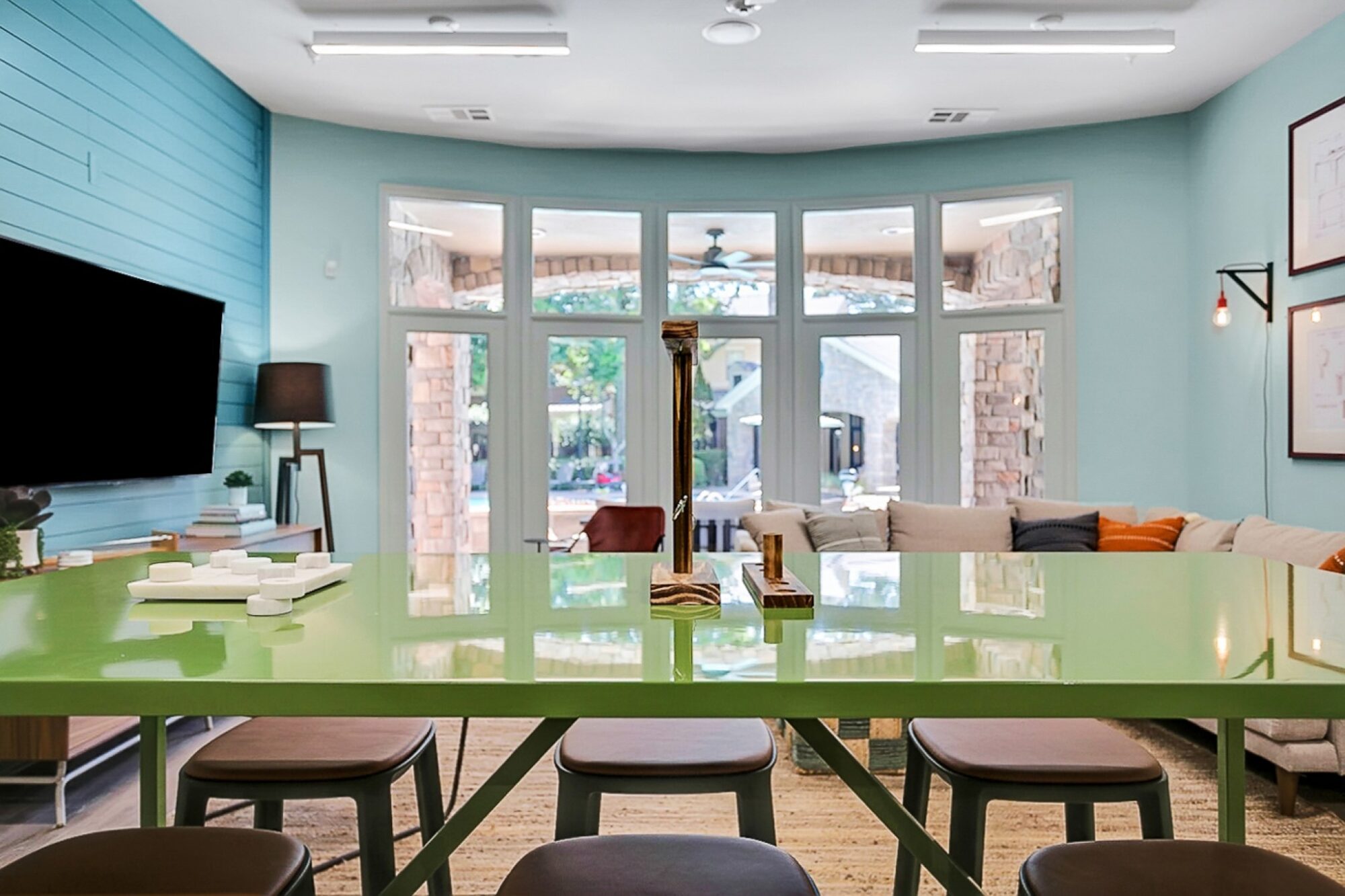 Modern living room with a large green table and stools, a cozy sectional sofa, wall-mounted TV, and large windows letting in natural light and showing an outdoor patio area.