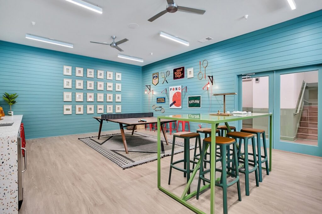 Modern game room with blue shiplap walls, tall green table with stools, wall art, ping pong table, ceiling fans, and light wood flooring. A door and stairs are visible on the right side.