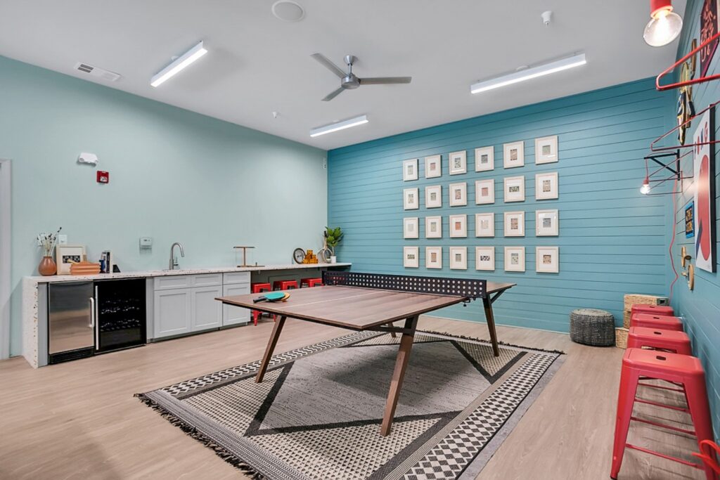 A modern game room with a ping pong table on a rug, a kitchenette with a sink and mini fridge, red stools at a counter, and framed artwork on a blue accent wall. Ceiling fans and bright lighting overhead.