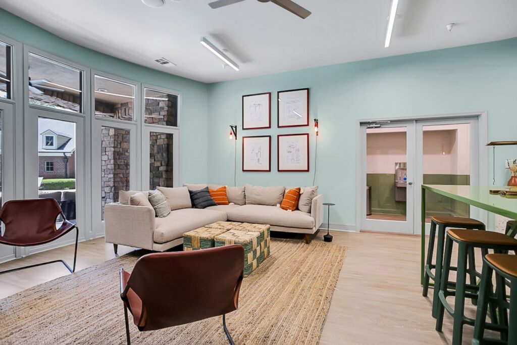 Modern living room with a light beige sectional sofa, patterned pillows, a woven rug, brown chairs, framed art on a pale blue wall, and a green breakfast bar with stools. Large windows let in natural light.