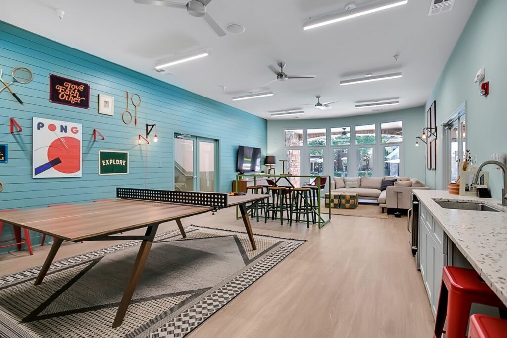 A bright, modern recreation room with a ping pong table, bar seating, a sofa, wall art, large windows, and teal accent walls. The space also features a kitchenette and light wood flooring.