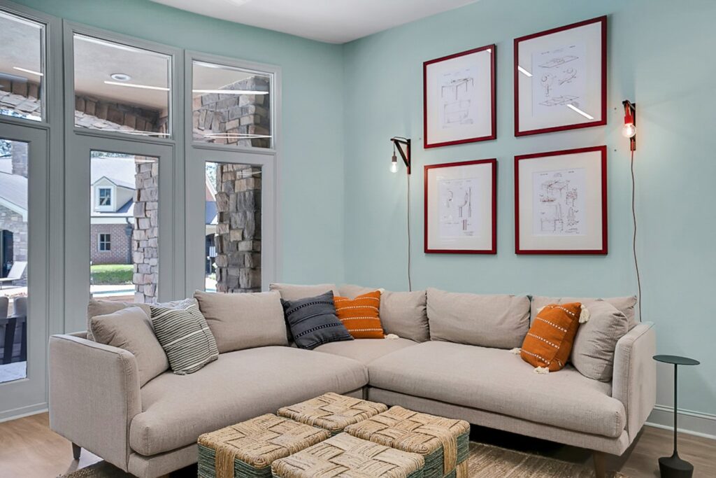 A modern living room with a light beige sectional sofa, assorted pillows, two woven ottomans, light blue walls, four framed art pieces, wall-mounted lights, and large windows overlooking a stone porch.