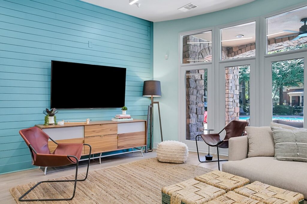 Modern living room with a light blue accent wall, wall-mounted TV, wooden console, two contemporary chairs, floor lamp, beige sofa, textured pouf, woven coffee tables, and large windows overlooking a patio and pool.