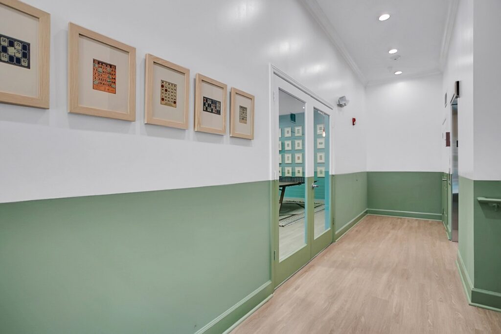 A hallway with light wood flooring and green lower walls, white upper walls, and framed artwork. Glass double doors at the end reveal a room with a table and matching framed art. Ceiling lights brighten the space.