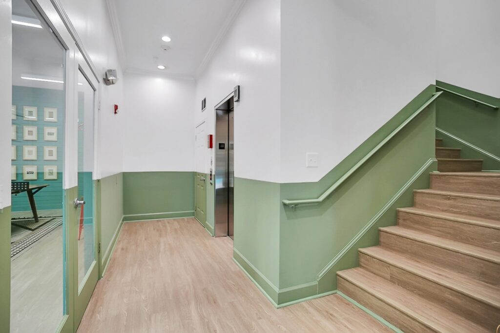 A clean hallway with light wood flooring, pale green and white walls, an elevator, a closed door, and a staircase with green trim ascending to the right. A glimpse of an adjacent room with wall art is visible through glass doors.