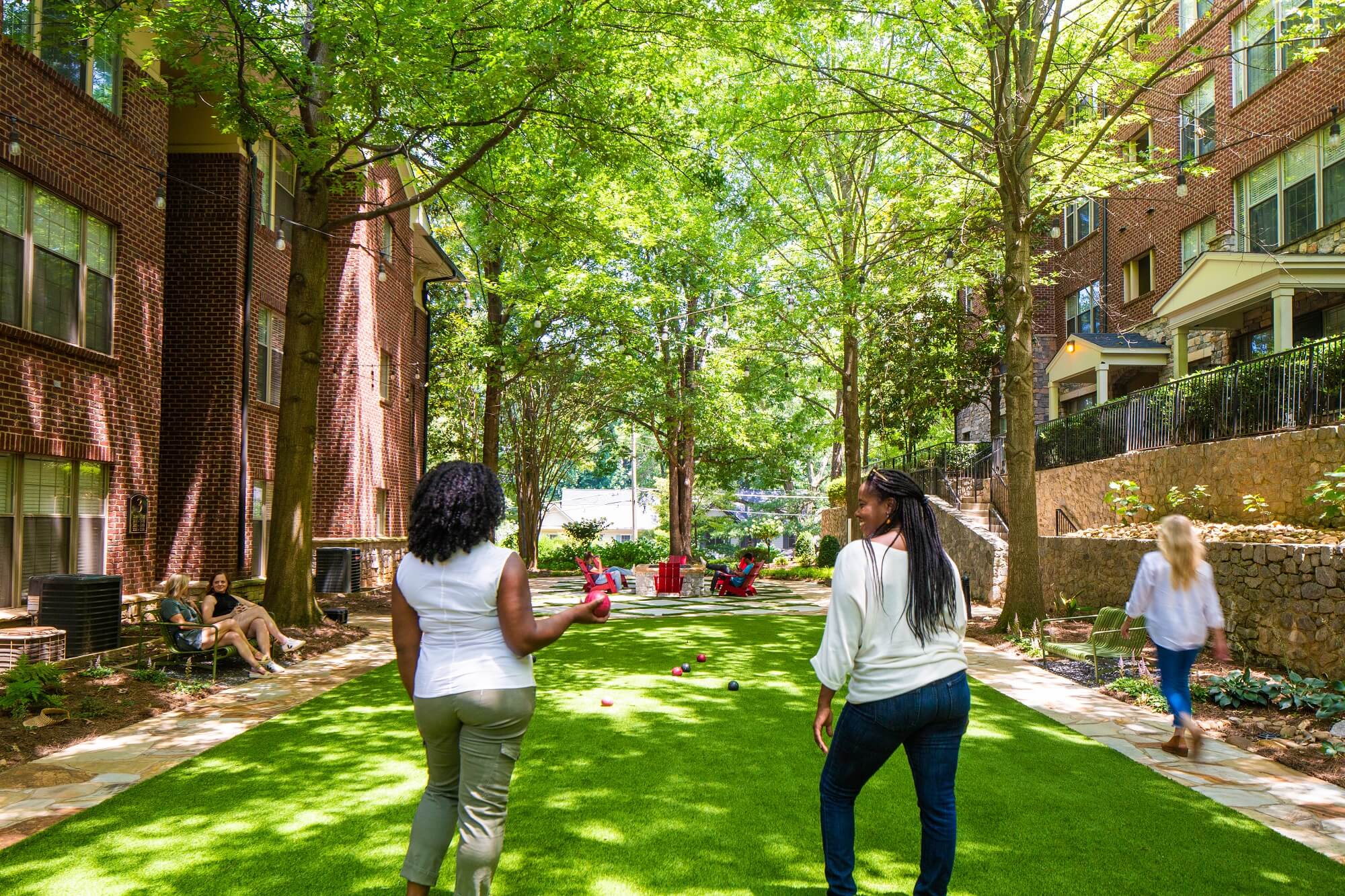 Two women walk across a green lawn surrounded by brick apartment buildings and trees, while people relax nearby on benches and red chairs in the shade.