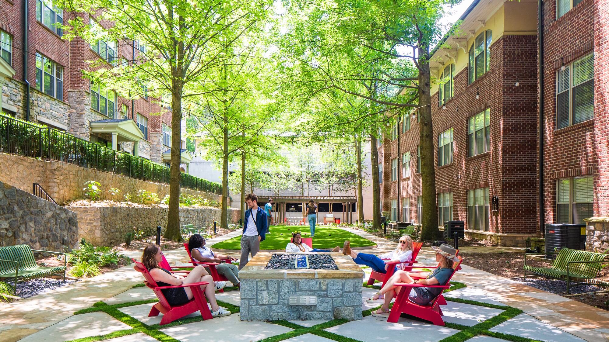 Five people relax in red chairs around a stone fire pit in a green, tree-filled courtyard between brick apartment buildings on a sunny day.