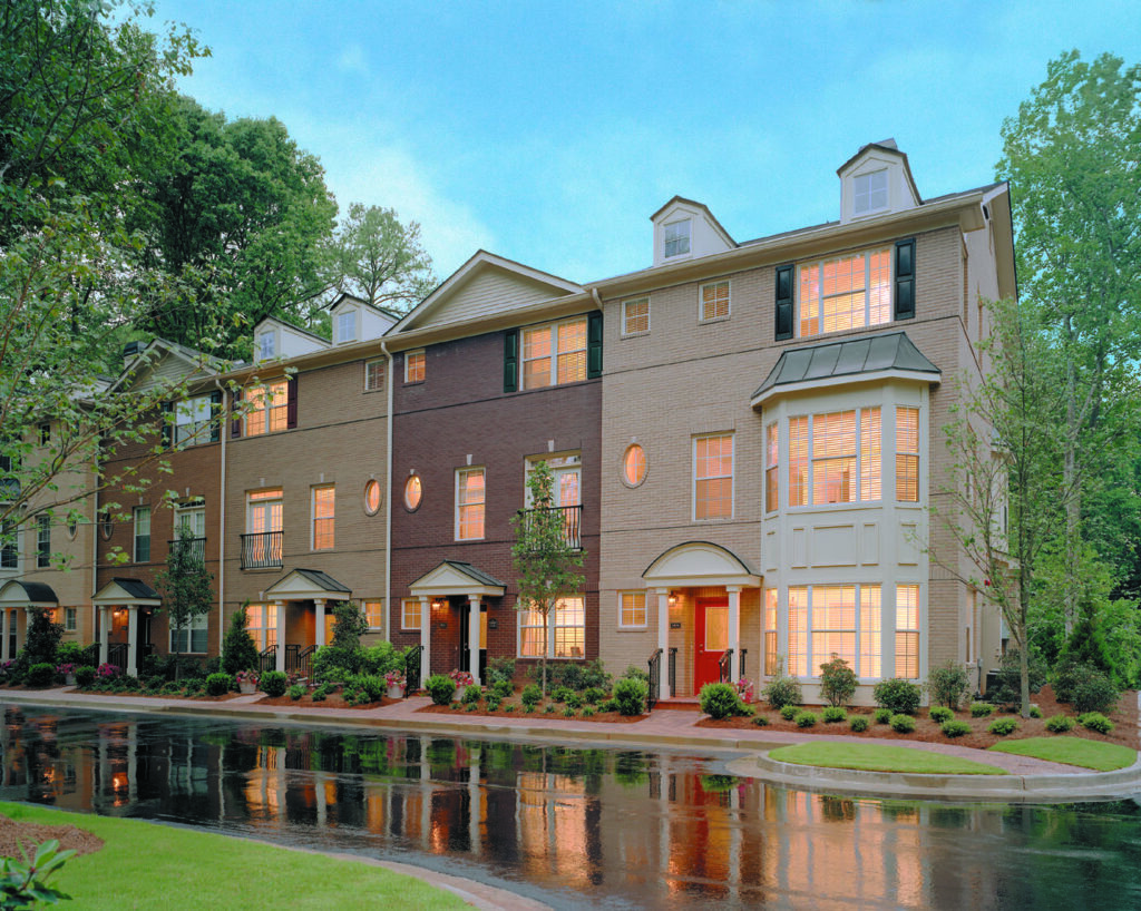 A row of three-story townhouses with lit windows on a wet street after rain, surrounded by trees and landscaped bushes under a bright sky.