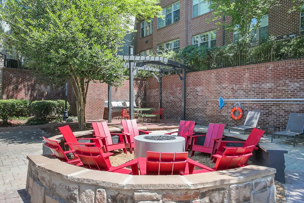 Outdoor patio with red Adirondack chairs arranged in a circle around a fire pit, set on a stone base. The area is surrounded by brick walls, greenery, and features a shaded pergola in the background.