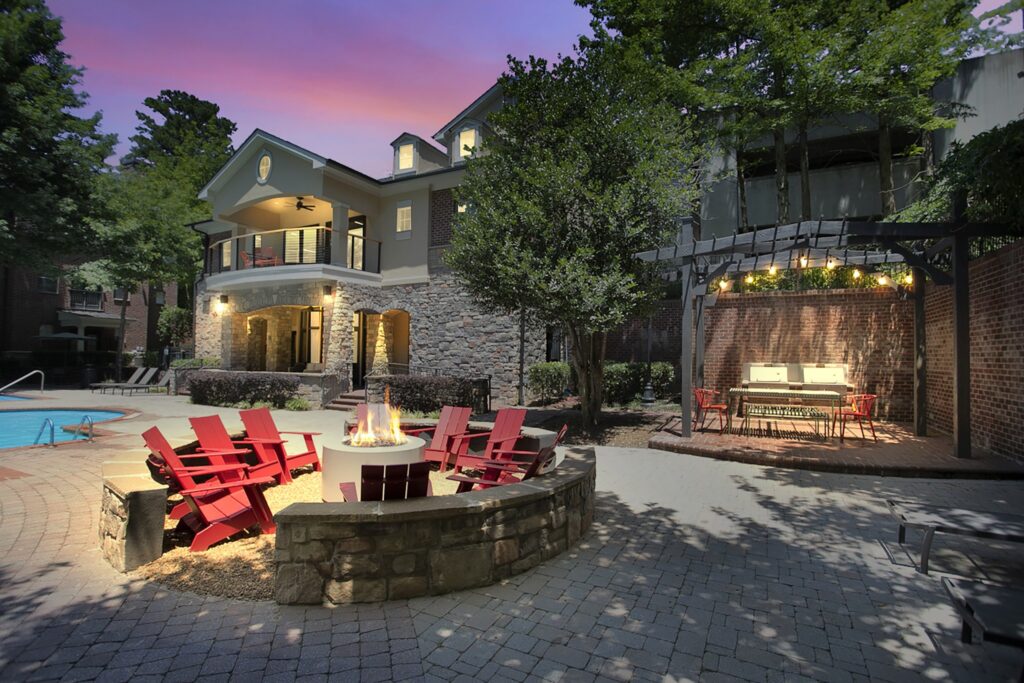 A cozy outdoor patio at dusk features red chairs around a fire pit, a grill under a pergola with string lights, and a large house with a pool in the background, surrounded by trees and stonework.