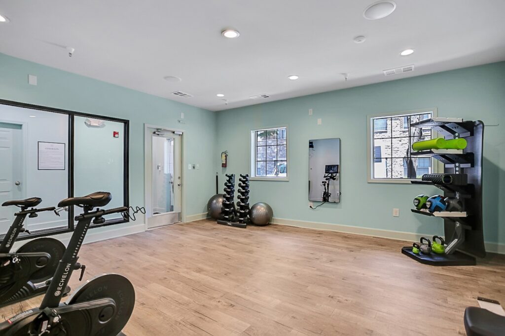 A modern fitness room with exercise bikes, free weights, stability balls, a mirror, and a storage rack holding mats and foam rollers. The room has light blue walls, wooden flooring, and windows letting in natural light.