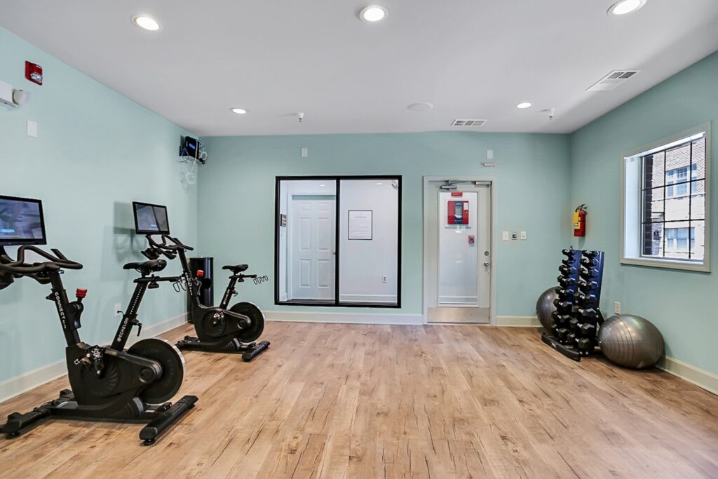 A fitness room with two stationary bikes, mounted screens, a rack of dumbbells, two exercise balls, wood-style flooring, and light blue-green walls. There are windows, a door, and overhead lighting.