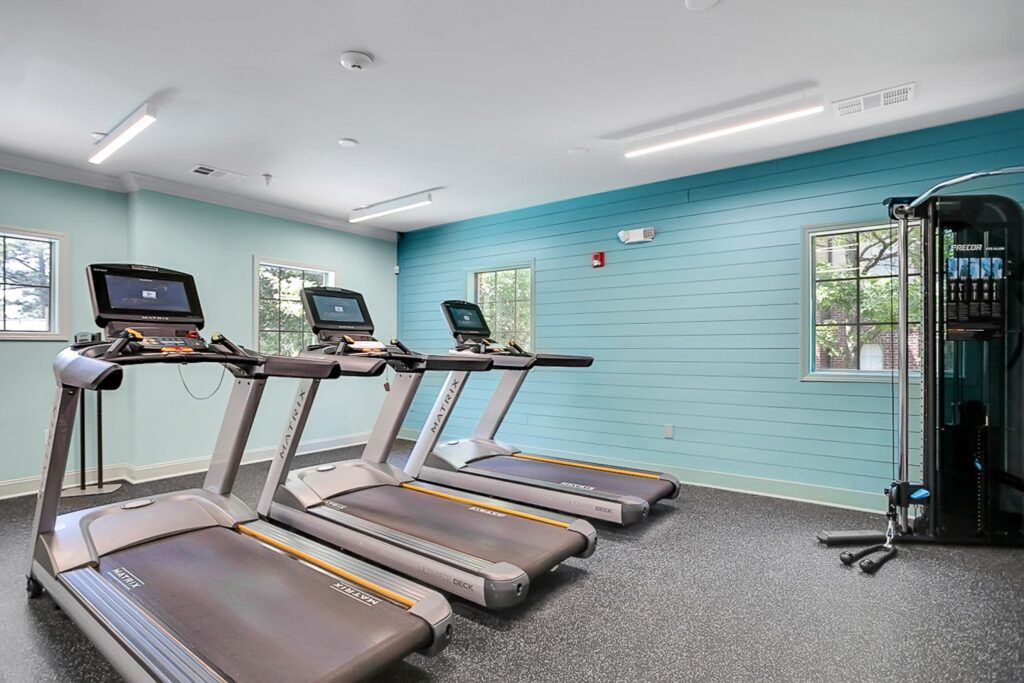 A modern gym with three treadmills side by side facing a wall, each with a built-in screen. The room has aqua walls, two windows, and a multi-use workout machine in the corner. The floor is covered with black speckled rubber.