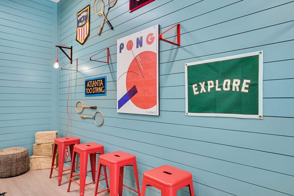 A wall with blue horizontal panels decorated with vintage sports-themed posters, two tennis rackets, and four red stools lined up beneath the artwork. Wicker baskets are on the floor in the corner.