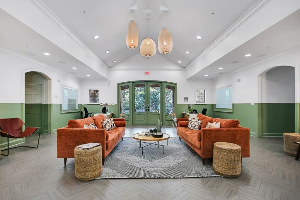 A modern lounge with orange sofas, round wicker ottomans, a gray rug, and a trio of woven pendant lights. Green double doors and white-green walls add a fresh, inviting feel; large windows let in natural light.