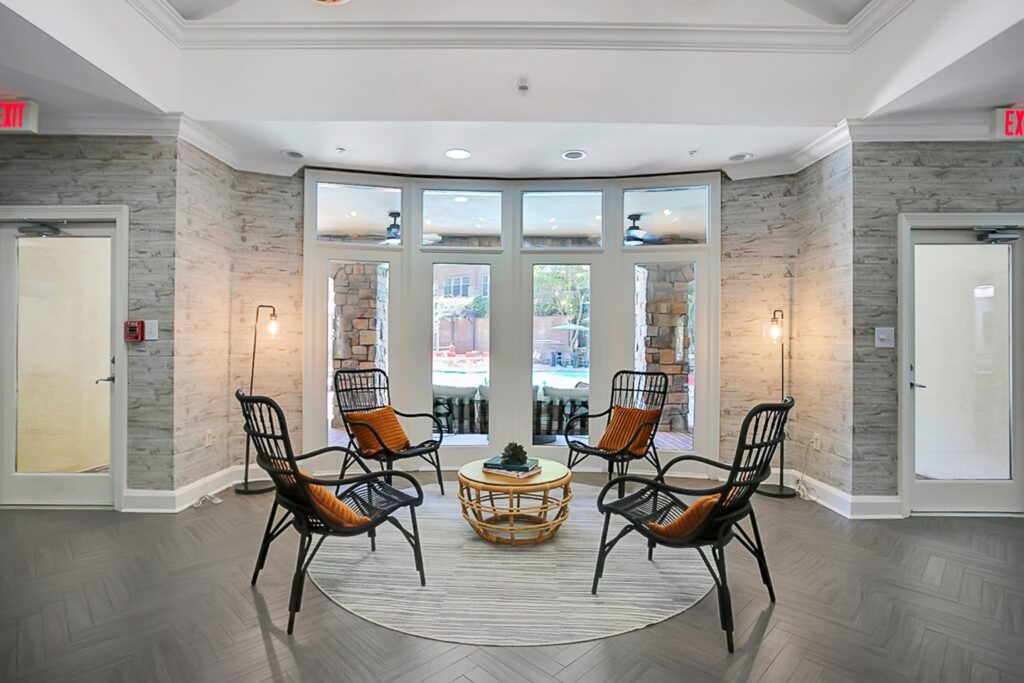 A modern lounge area with four black chairs arranged around a round wicker table on a circular rug, large windows in the background, and soft lighting from two floor lamps.