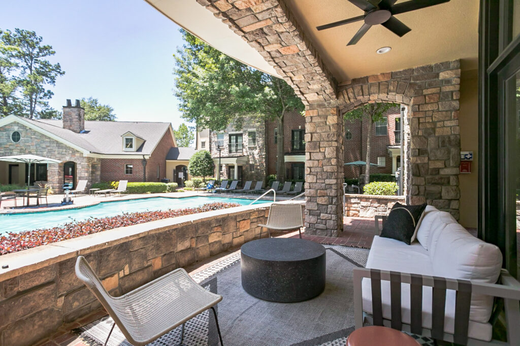 A shaded stone patio with modern seating overlooks a swimming pool surrounded by lounge chairs and brick buildings on a sunny day.