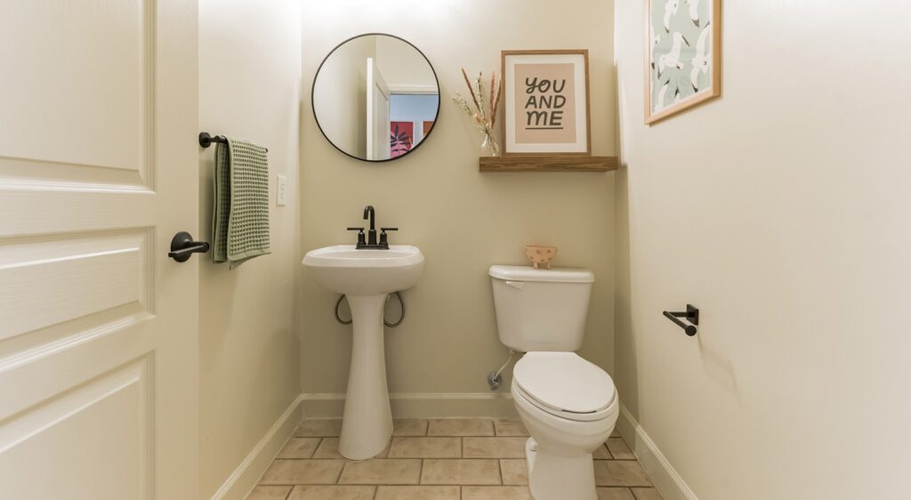 A small, modern bathroom with a pedestal sink, round mirror, white toilet, towel on a rack, and framed art above a floating shelf with a small plant. The walls are light-colored and the floor is tiled.