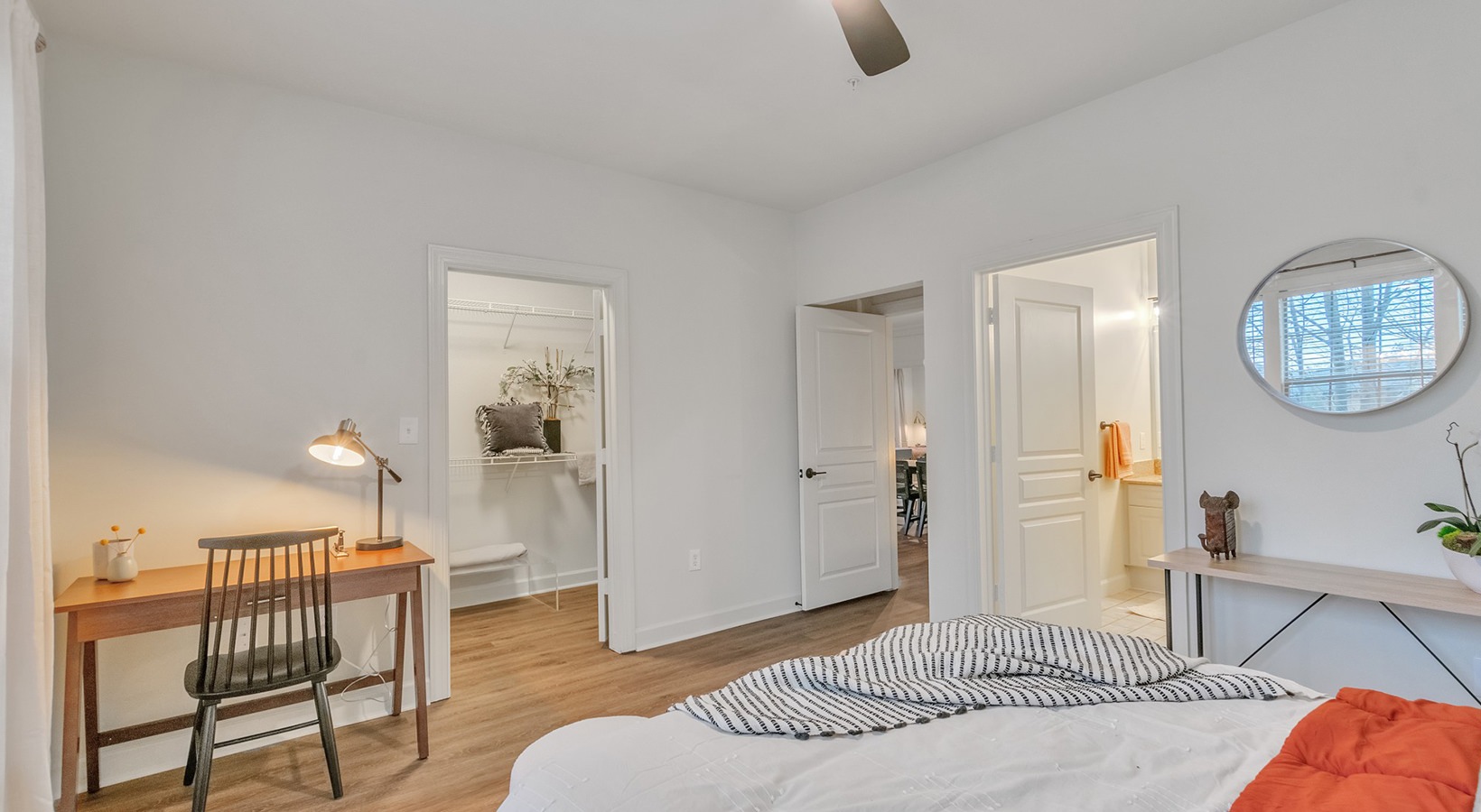 A bright bedroom with a neatly made bed, a wooden desk with a chair and lamp, a walk-in closet, an open bathroom door, and a round mirror above a side table with decor. Light wood floors and white walls.
