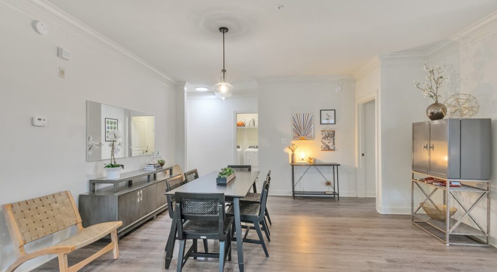 A modern dining room with a black dining table and six chairs, light wooden floors, neutral walls, and contemporary decor. A pendant light hangs above the table, and there is minimalist artwork and furniture throughout the space.