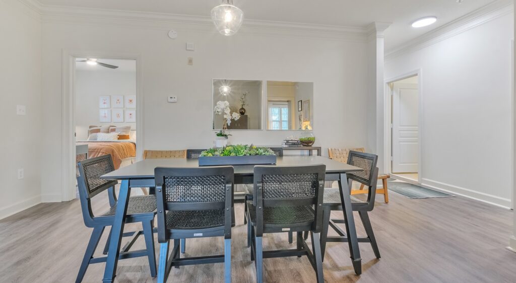 A modern dining room with a black table and six black chairs, a large wall mirror, indoor plants as centerpiece, and a view of a bedroom through an open doorway. The room has light walls and wood flooring.