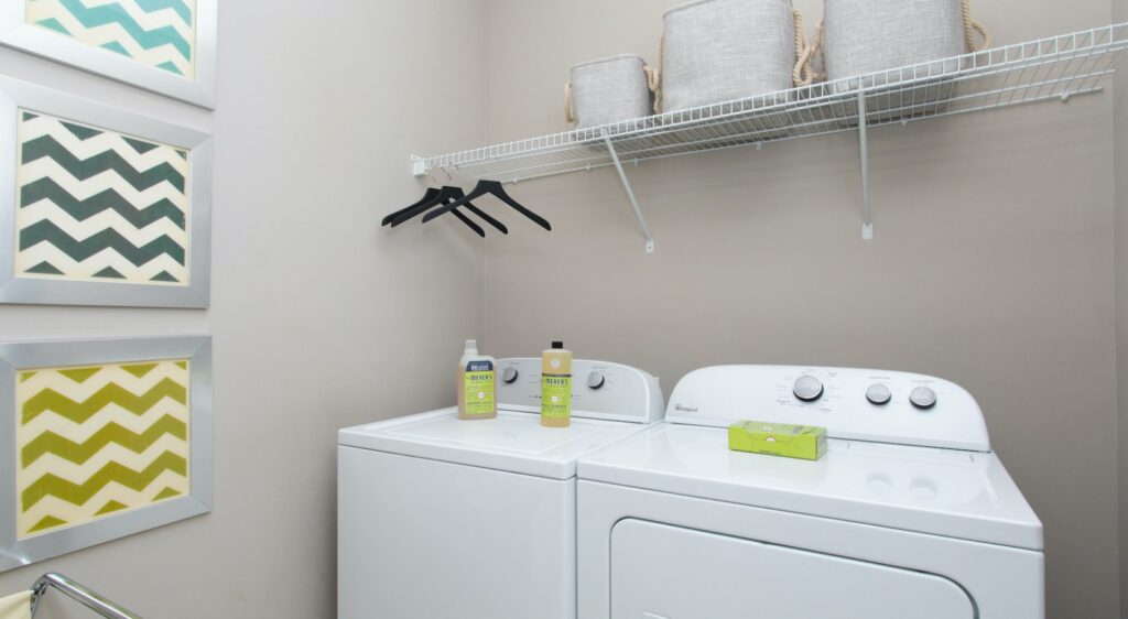 A laundry room with a white washer and dryer, detergent bottles on top, black hangers on a shelf, storage baskets above, and framed chevron-patterned artwork on the beige wall.