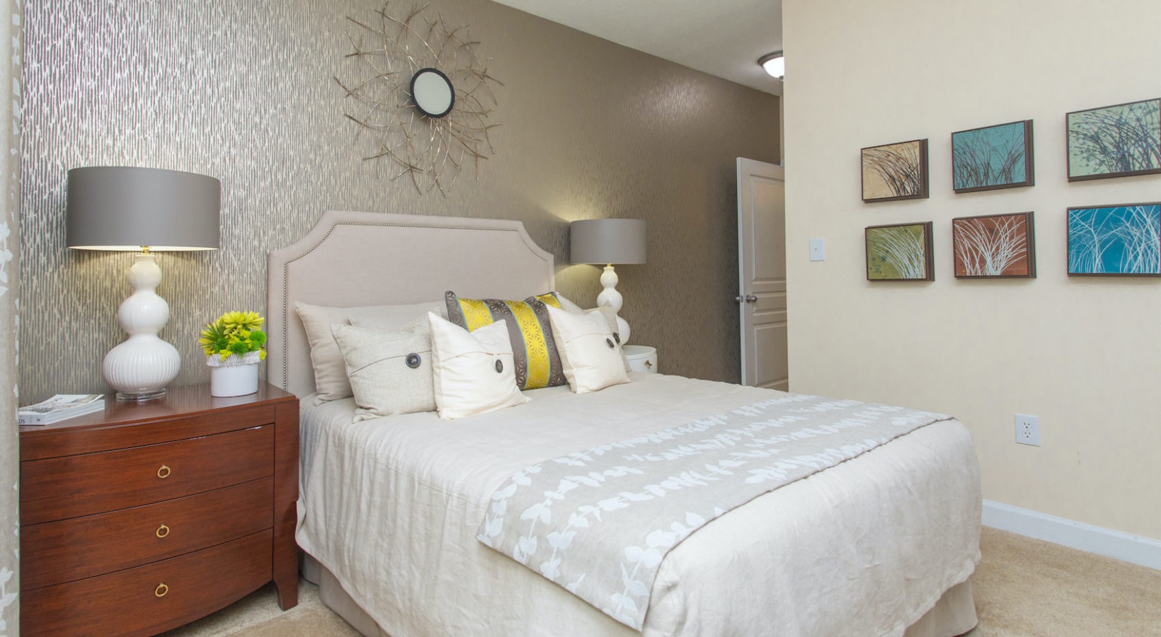A modern bedroom with a beige bed, decorative pillows, two white lamps on wooden nightstands, silver textured wallpaper, abstract wall art, and a sunburst mirror above the bed.