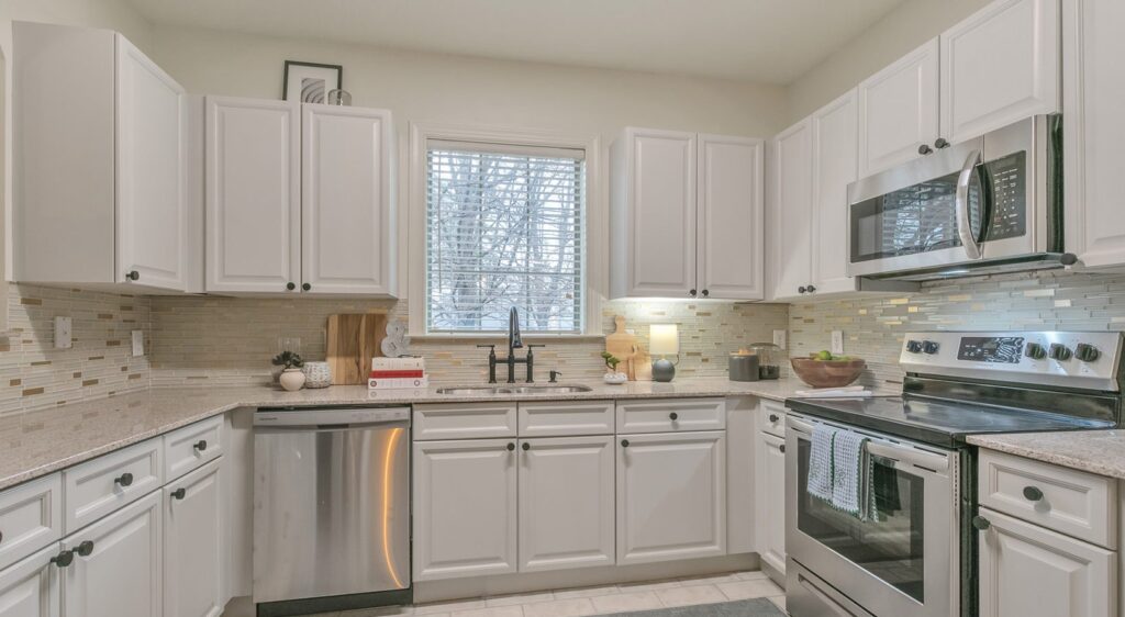 Modern kitchen with white cabinets, stainless steel appliances, a window with blinds above the sink, a light tile backsplash, and neutral countertops. A dishwasher, stove, and microwave are visible.
