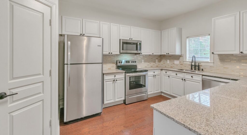Modern kitchen with white cabinets, stainless steel appliances, granite countertops, tile backsplash, and wood flooring. A window above the sink brings in natural light.