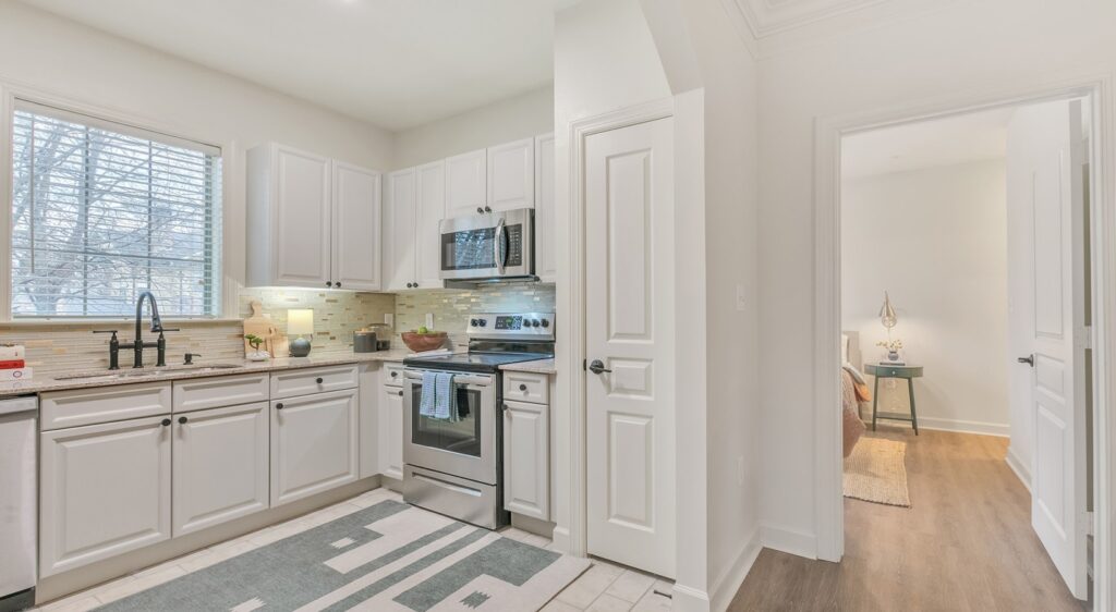 A bright, modern kitchen with white cabinets, stainless steel appliances, and a tiled backsplash. There is a window above the sink, a patterned rug on the floor, and an open door leading to a cozy room with a small table.