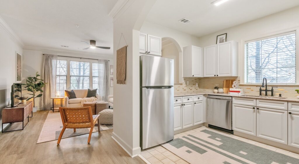 Modern open-concept living room and kitchen, featuring white cabinets, stainless steel appliances, light wood flooring, a cozy sitting area with a sofa and chair, and large windows letting in natural light.