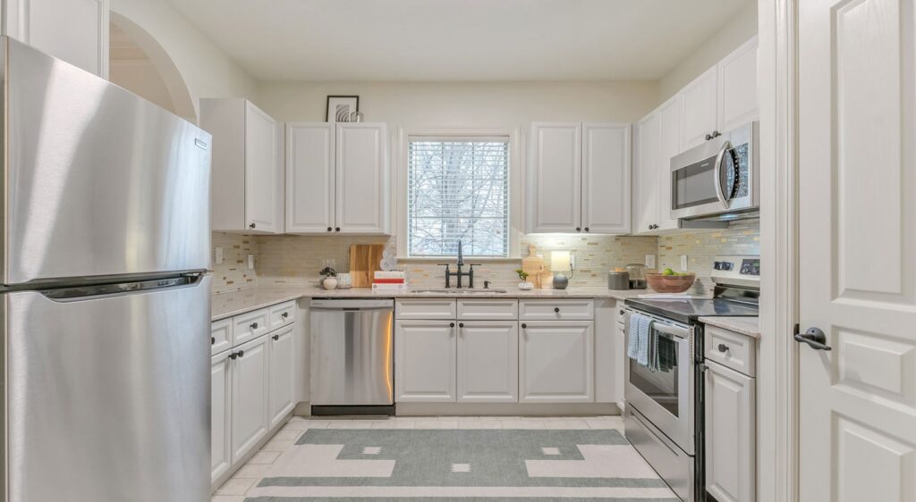 Modern kitchen with white cabinets, stainless steel appliances, a window above the sink, light tile backsplash, and a patterned rug on the floor. Countertops hold various kitchen items and decor.