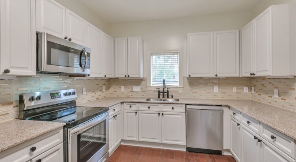 Modern kitchen with white cabinets, stainless steel appliances, granite countertops, beige mosaic tile backsplash, and a small window above the sink. The kitchen has wooden floors and under-cabinet lighting.