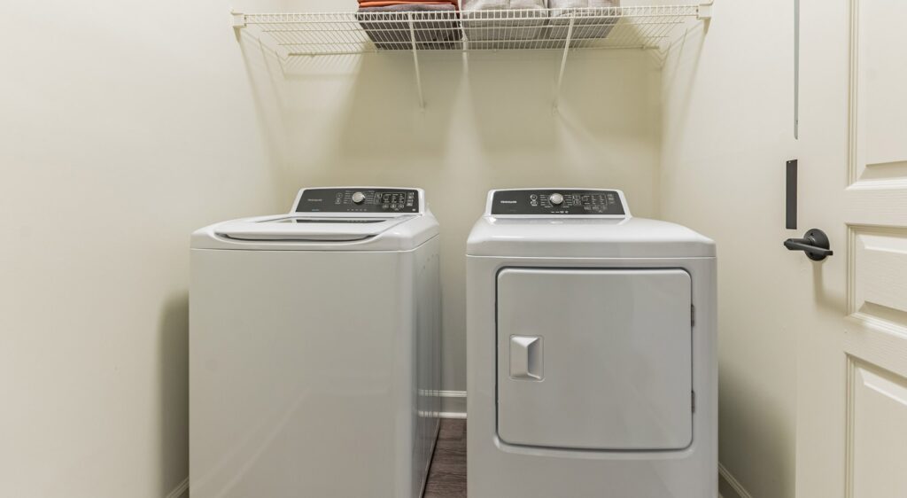 A laundry room with a white top-loading washing machine and a matching dryer side by side, beneath a wire shelf holding folded towels against a light-colored wall.