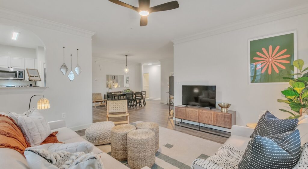 Bright living room with a modern sofa, two woven ottomans, a TV on a wooden stand, and a large art print of an orange flower. Open layout leads to a dining area and white kitchen. Neutral tones and cozy decor throughout.