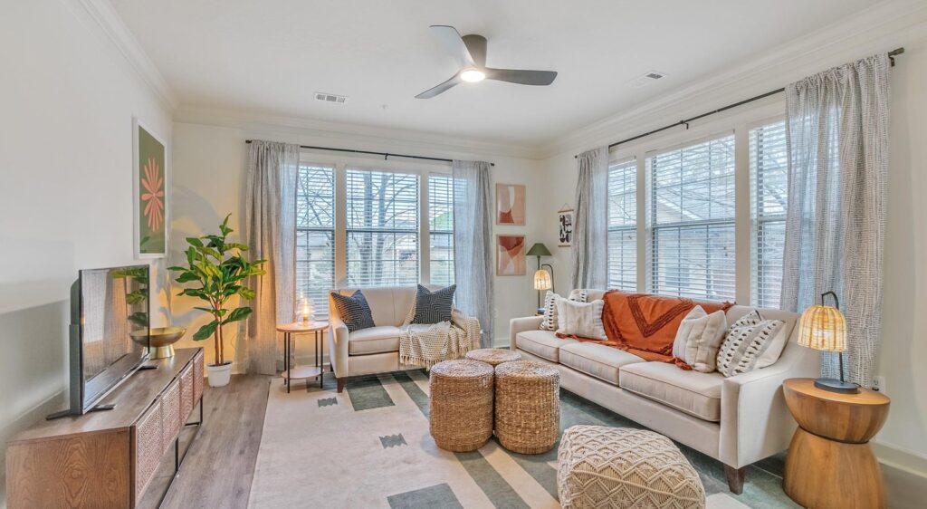 Bright living room with large windows, a white sofa, an armchair, two woven ottomans, a TV on a wooden stand, a potted plant, decorative pillows, and modern decor in neutral and earthy tones.