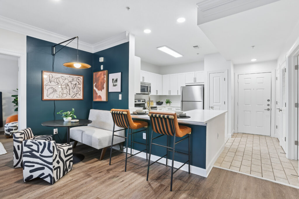 Modern apartment kitchen with white cabinets, stainless steel appliances, a breakfast bar with two orange stools, and a small dining nook with abstract art and zebra-print chairs. Wood and tile flooring throughout.