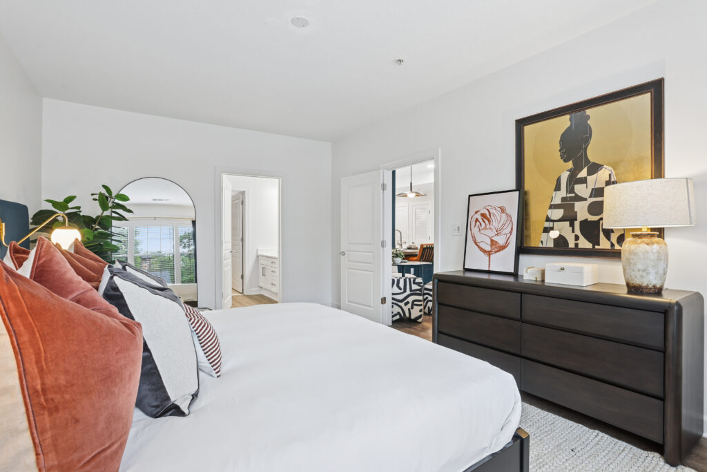 Modern bedroom with a neatly made bed, rust and black accent pillows, dark dresser, large artwork, table lamp, mirror, houseplant, and open doors leading to a bathroom and another room.
