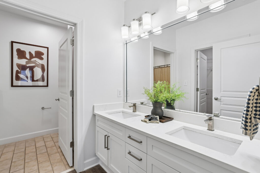 Modern bathroom with a double sink vanity, large mirror, white cabinets, silver fixtures, potted plant, and wall art. A door leads to a toilet area with tiled floor and a towel hanging on the vanity.