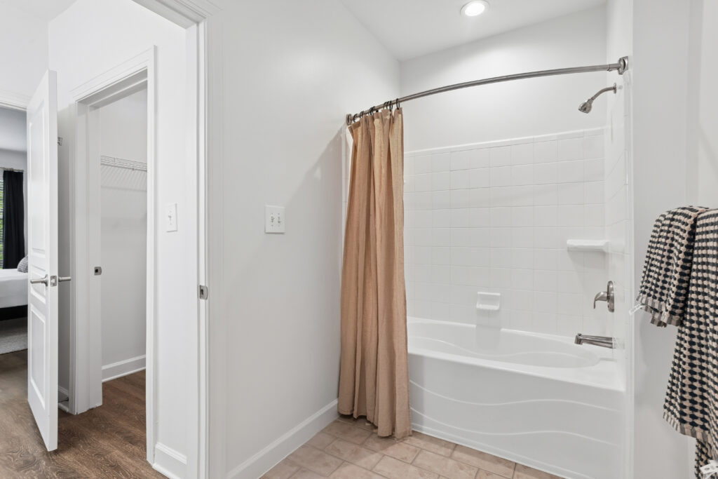A modern bathroom with a white bathtub and tile surround, a beige shower curtain, towel on the wall, and two open doors leading to a closet and a bedroom with wood flooring.