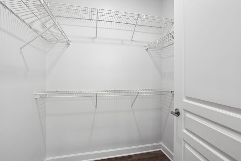 Empty walk-in closet with white wire shelving on three sides, white walls, and a white door slightly open. The floor is dark wood. The space is clean and well-lit.
