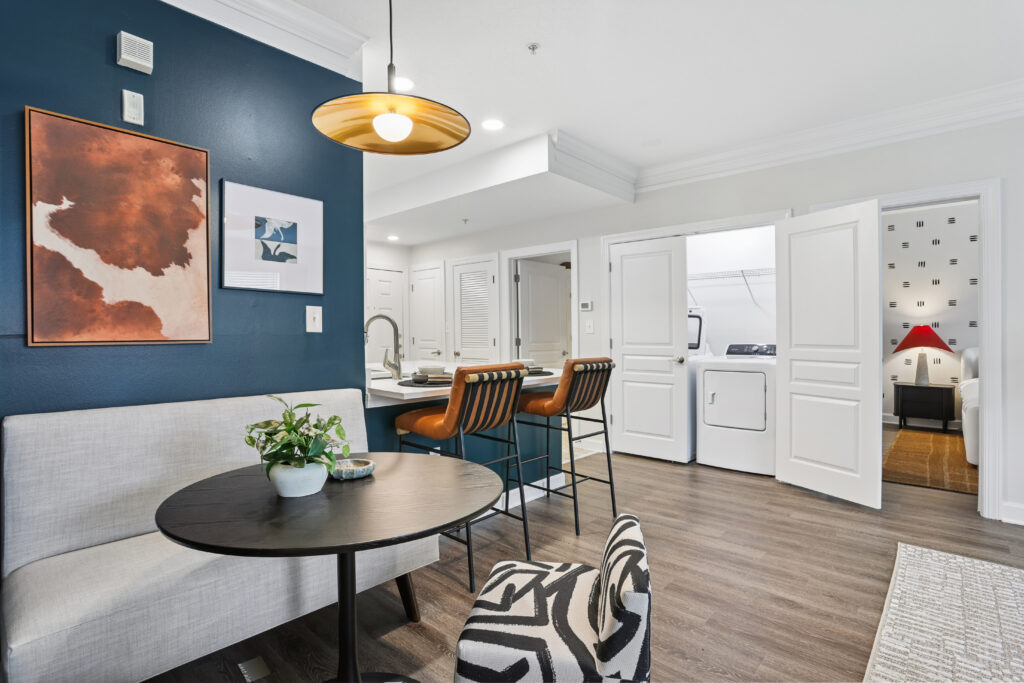 Modern open-plan living space with a dark blue accent wall, round dining table, built-in bench, two barstools at a kitchen island, laundry area with washer and dryer, and a glimpse of a cozy room with a red lamp.