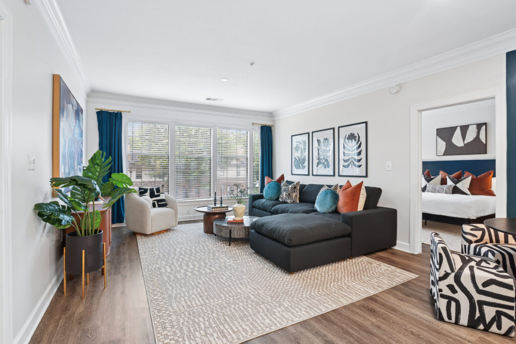 Modern living room with a large gray sectional sofa, patterned armchair, potted plant, abstract wall art, and a textured rug. Large windows with blinds let in natural light. A bedroom is visible through an open doorway.