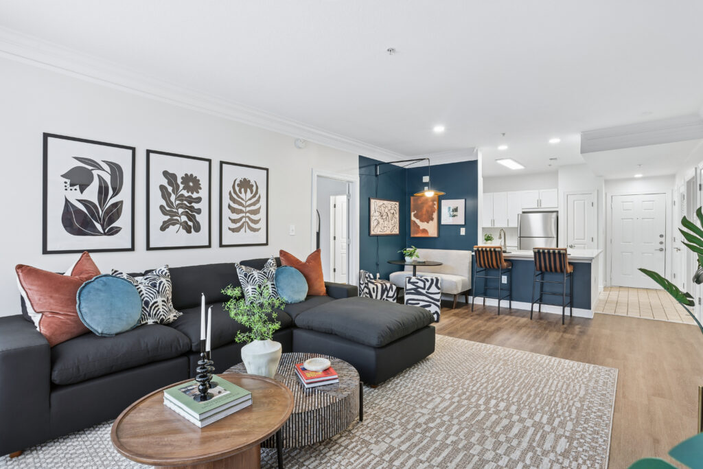 Modern open-concept living room and kitchen with a dark sectional sofa, patterned pillows, a round coffee table, framed botanical art, blue accent wall, bar stools, and wood flooring.