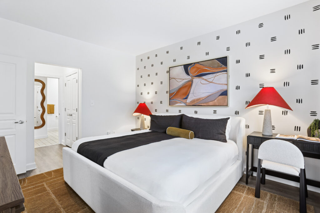 A modern bedroom with a white bed, black and white bedding, two red-shaded lamps on side tables, abstract wall art, a desk with a chair, and a view into a bathroom through an open door.