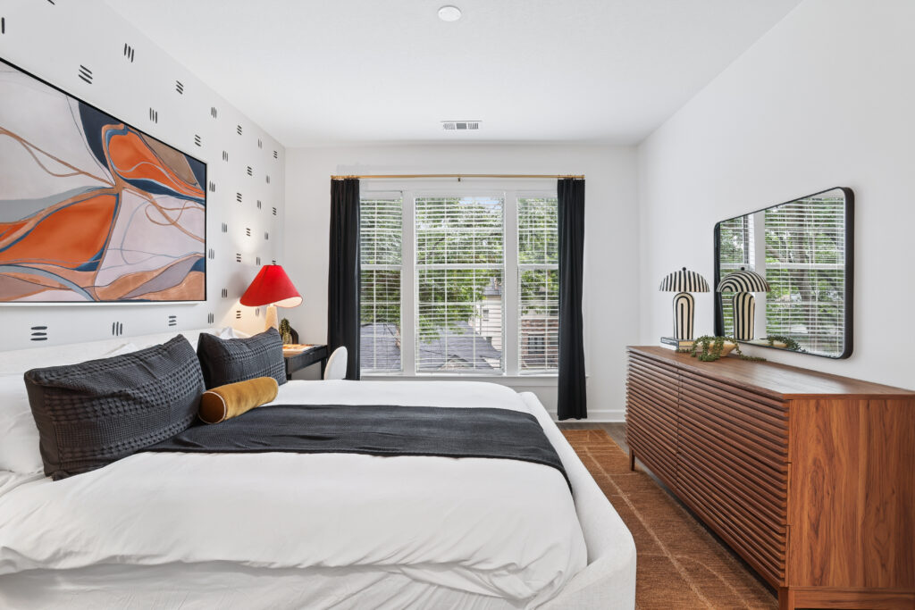 Modern bedroom with a white bed, black and gold pillows, abstract wall art, wooden dresser with decor, desk with red lamp, and large window with black curtains letting in natural light.