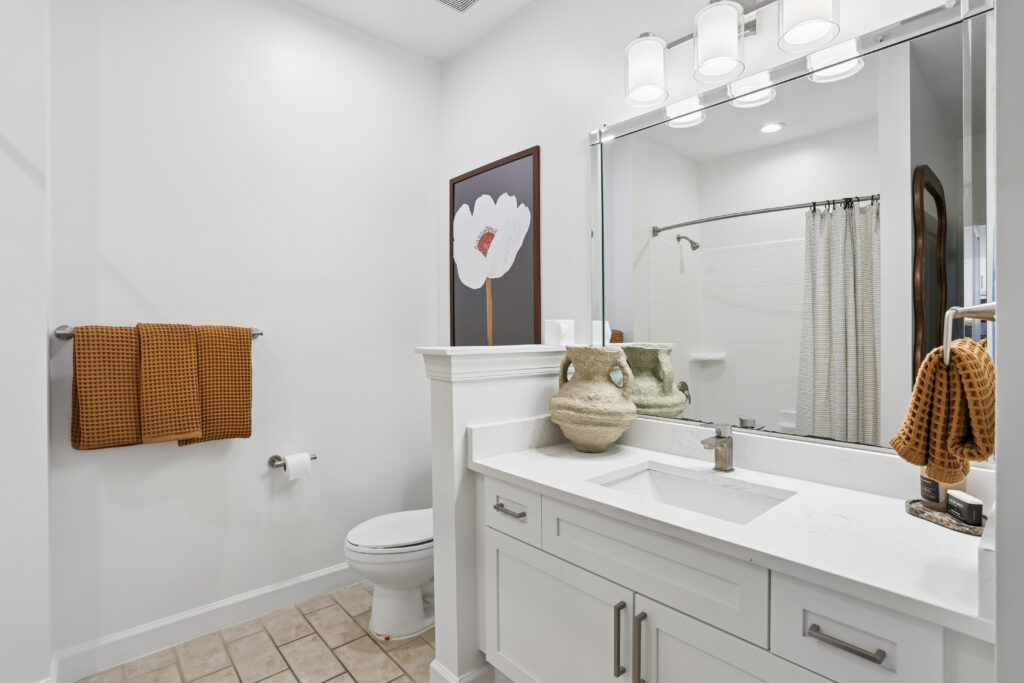 Modern bathroom with white walls and cabinetry, tan tiled floor, a white countertop with decorative vases, a large mirror, brown towels, a wall art of a flower, and a shower with a white curtain.